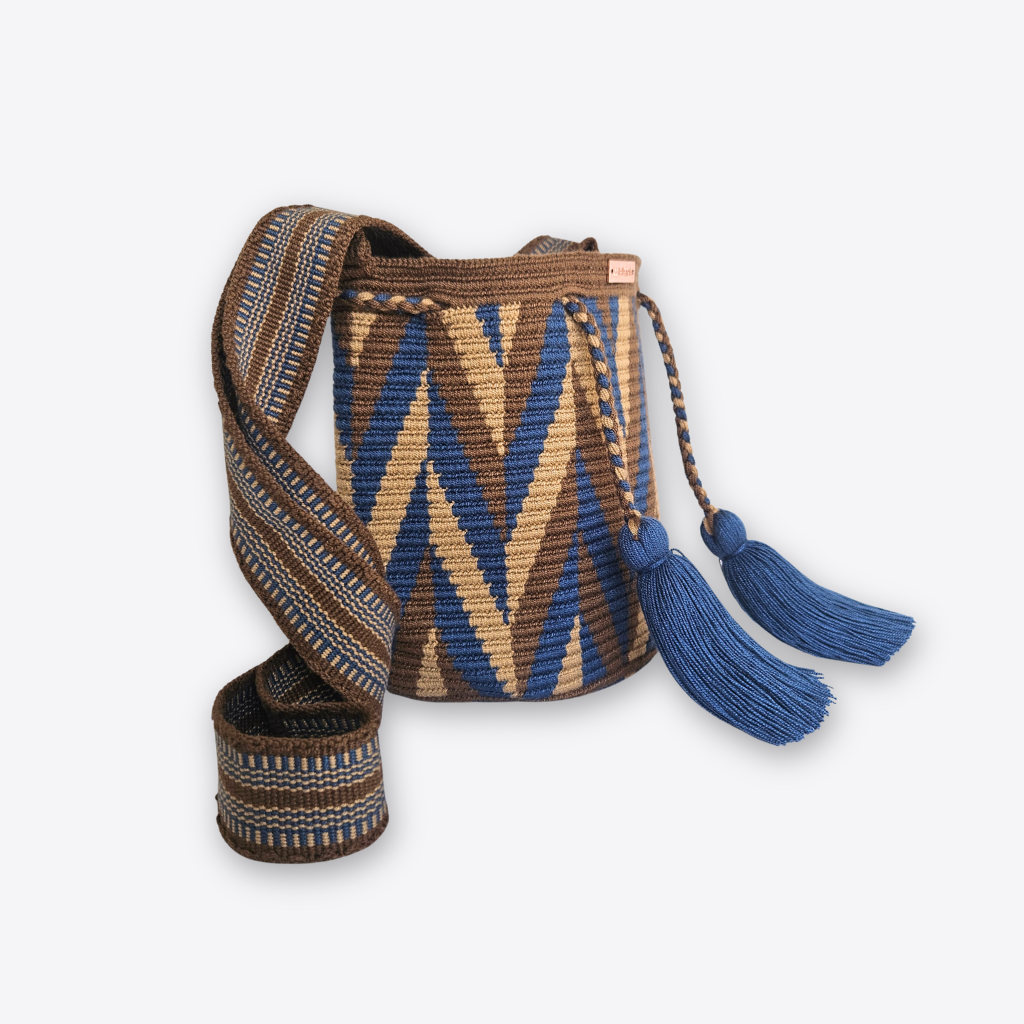 A handwoven bucket bag with a geometric chevron pattern in blue, brown, and beige, featuring a matching woven shoulder strap and two blue tassels, shown on a light gray background.