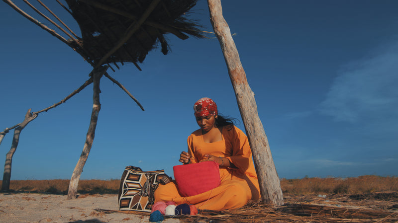 Wayuu woman  in orange outfit sitting under a tree with a red bag, against a clear blue sky.